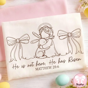 Puede incluir: Tela blanca rota con temática de Pascua con un dibujo lineal marrón de una figura abrazando a un conejito, con dos lazos a cada lado. El texto "He is not here, He has Risen" y "MATTHEW 28:6" están impresos debajo.