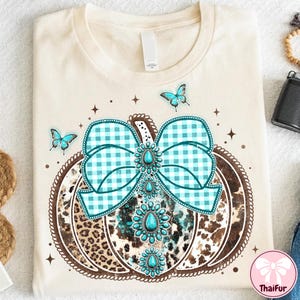 May include: Cream-colored t-shirt featuring a pumpkin design with a turquoise and blue gingham bow, turquoise gemstones, and a leopard print pattern. The pumpkin is outlined in brown with a decorative border, and includes three blue butterflies.