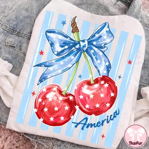 May include: White pet shirt featuring a patriotic cherry design. The cherries are red with white stars, and a blue bow with white stars sits above them. The word "America" is written in blue script below the cherries. Blue and white striped background.