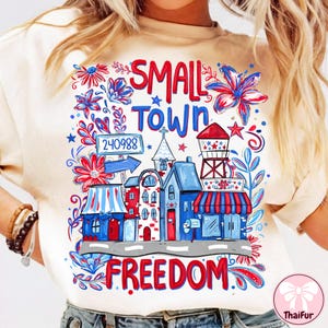 May include: Cream-colored t-shirt featuring a colorful graphic design. The design includes the words "SMALL TOWN FREEDOM" in red, along with illustrations of buildings, flowers, and a water tower in red, white, and blue. The zip code 240988 is also included.