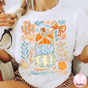 May include: White t-shirt featuring a colorful pumpkin design with floral and bow accents. The pumpkin is orange and blue, surrounded by orange, blue, and yellow floral elements. The design is centered on the front of the shirt.
