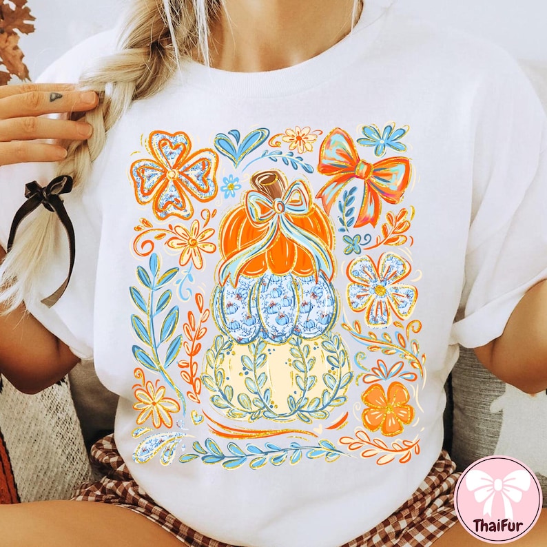 May include: White t-shirt featuring a colorful pumpkin design with floral and bow accents. The pumpkin is orange and blue, surrounded by orange, blue, and yellow floral elements. The design is centered on the front of the shirt.