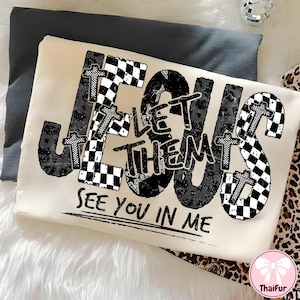 May include: Cream-colored sweatshirt with the words "JESUS LET THEM SEE YOU IN ME" in a black and white design. The word "JESUS" features checkered and leopard print patterns. The sweatshirt is folded on a gray surface.