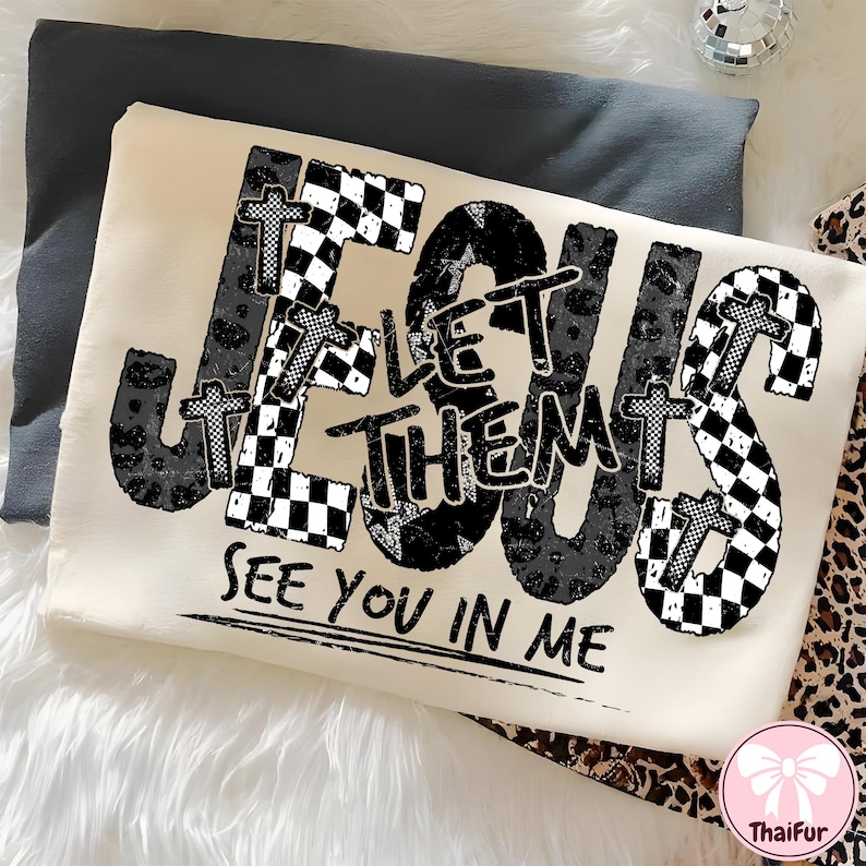 May include: Cream-colored sweatshirt with the words "JESUS LET THEM SEE YOU IN ME" in a black and white design. The word "JESUS" features checkered and leopard print patterns. The sweatshirt is folded on a gray surface.