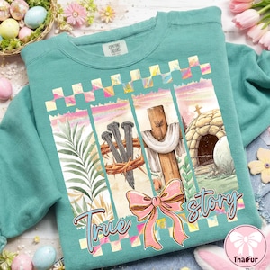 May include: Teal sweatshirt with a colorful graphic featuring a cross, nails, a crown of thorns, and a pink bow. The words "True Story" are written on the front. The sweatshirt is a comfortable, casual style.
