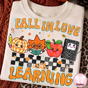 May include: A cream-colored t-shirt featuring a graphic with the text "FALL IN LOVE WITH LEARNING" in orange. The design includes a cartoon pumpkin, a leaf with glasses reading a book, an apple with a worm, and a notebook on a checkered path.