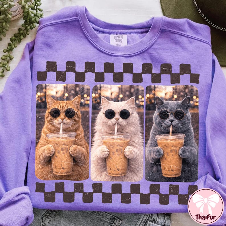 May include: A purple Comfort Colors sweatshirt featuring a graphic of three cats wearing sunglasses and drinking iced coffee. The cats are in individual frames with a brown checkered border. The sweatshirt has a crew neck.