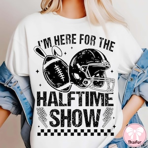 Può includere: T-shirt bianca con un design grafico nero raffigurante un pallone da football americano, un casco e la scritta "I'M HERE FOR THE HALFTIME SHOW". Il design include stelle e un motivo a scacchi.
