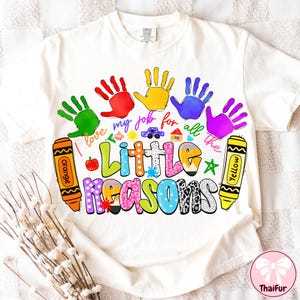 May include: Cream-colored t-shirt with a colorful graphic design. The design includes handprints, crayons, and the text "I love my job for all the Little Reasons." A cute car and house are also included in the design.