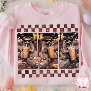 Puede incluir: Sudadera rosa claro con un diseño de tres mapaches con gafas de sol bebiendo café helado. Los mapaches están enmarcados dentro de un borde a cuadros marrón. La sudadera tiene mangas largas y cuello redondo.