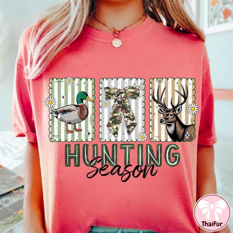 Puede incluir: Camiseta de color coral con tres ilustraciones enmarcadas: un pato, un lazo de camuflaje y un ciervo. Las palabras "HUNTING Season" est&aacute;n impresas debajo en verde y blanco. El dise&ntilde;o tiene un fondo a rayas.
