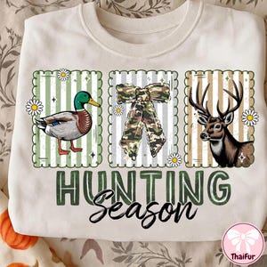 Puede incluir: Sudadera crema con tem&aacute;tica de caza. Presenta un pato, un lazo de camuflaje y un ciervo, cada uno en un dise&ntilde;o enmarcado. Las palabras "HUNTING Season" est&aacute;n impresas en verde y negro. Se incluyen acentos de margaritas.