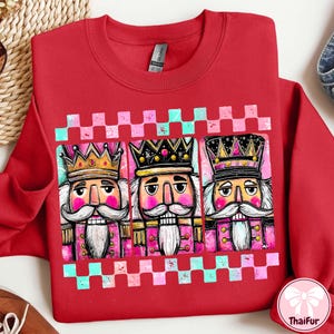 K&ouml;nnte beinhalten: Ein rotes Sweatshirt mit einer Grafik von drei Nussknacker-Figuren mit Kronen und rosa und wei&szlig;en Akzenten. Das Design wird von einem rosa-t&uuml;rkisfarbenen Schachbrettmuster umrahmt.