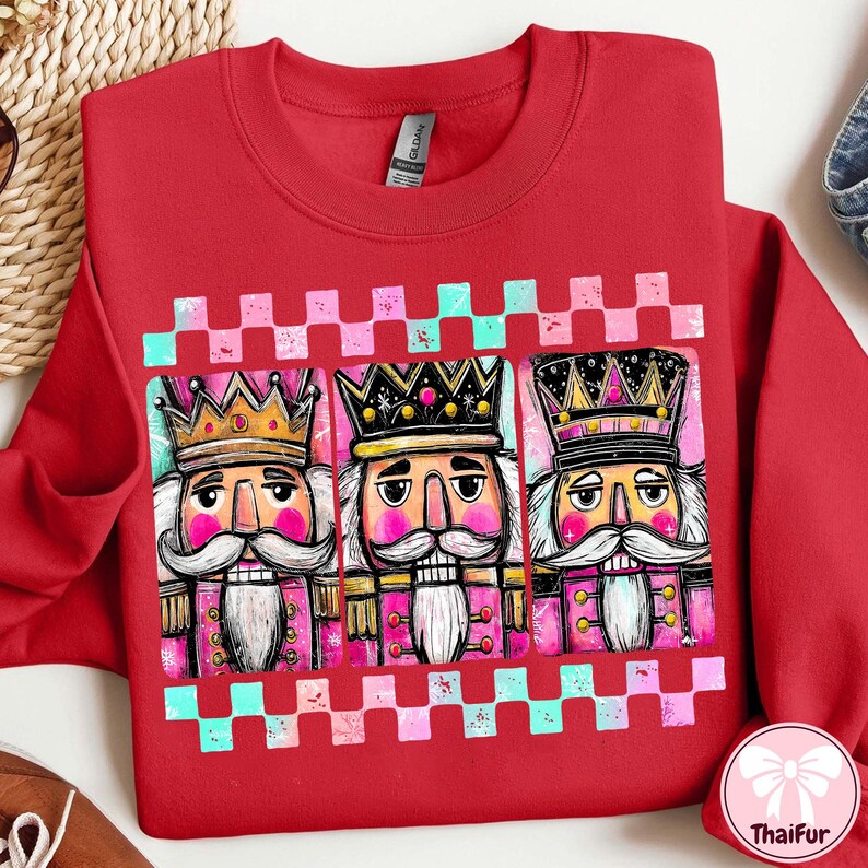 K&ouml;nnte beinhalten: Ein rotes Sweatshirt mit einer Grafik von drei Nussknacker-Figuren mit Kronen und rosa und wei&szlig;en Akzenten. Das Design wird von einem rosa-t&uuml;rkisfarbenen Schachbrettmuster umrahmt.