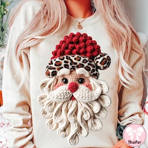 May include: A cream-coloured sweatshirt with a Santa Claus design. The Santa face is made with textured yarn, featuring a red nose, white beard, and a leopard print hat. The design is centered on the front of the garment.