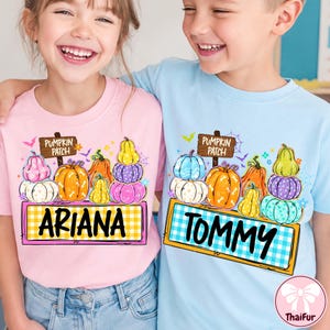 Può includere: Una t-shirt rosa chiaro con una grafica di zucche impilate e il testo 'ARIANA' e 'PUMPKIN PATCH', e una t-shirt azzurra con una grafica di zucche impilate e il testo 'TOMMY' e 'PUMPKIN PATCH' stampati sulla parte anteriore.