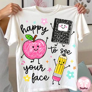 以下が含まれることがあります： 白い半袖Tシャツで、ピンクのリンゴ、黒いノート、顔の描かれた黄色い鉛筆のグラフィックプリントが施されています。周囲にはカラフルな星や花が散りばめられ、黒い文字で「happy to see your face」と書かれています。