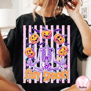 May include: A black t-shirt featuring a graphic of a purple skeleton sitting in a lotus position surrounded by orange jack-o'-lanterns, small purple bats, and yellow stars against a pink and black striped background with the text Stay Spooky.