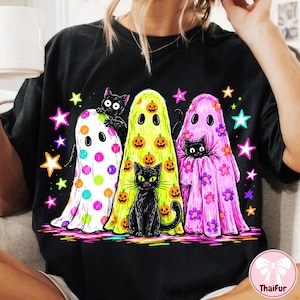 Pu&ograve; includere: Una t-shirt nera con una stampa grafica di tre fantasmi e due gatti neri. I fantasmi sono bianchi con pois colorati, gialli con zucche arancioni e rosa con fiori viola. Stelle dai colori vivaci circondano le figure sul capo di abbigliamento.