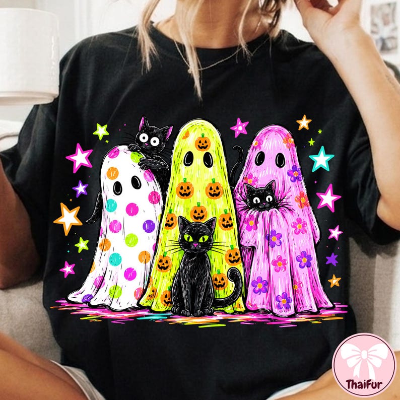 Pu&ograve; includere: Una t-shirt nera con una stampa grafica di tre fantasmi e due gatti neri. I fantasmi sono bianchi con pois colorati, gialli con zucche arancioni e rosa con fiori viola. Stelle dai colori vivaci circondano le figure sul capo di abbigliamento.