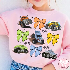 K&ouml;nnte beinhalten: Hellrosa Sweatshirt mit bunten Monstertruck-Grafiken und dekorativen Schleifen. Die Monstertrucks sind orange, gelb, gr&uuml;n und blau. Die Schleifen sind orange, gr&uuml;n, gelb und blau.