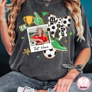 May include: A gray t-shirt with a soccer-themed design. The design includes a soccer ball, a trophy, a soccer cleat, a bow with the number "01" and the word "NAME", and a photo frame with the text "Text Here".