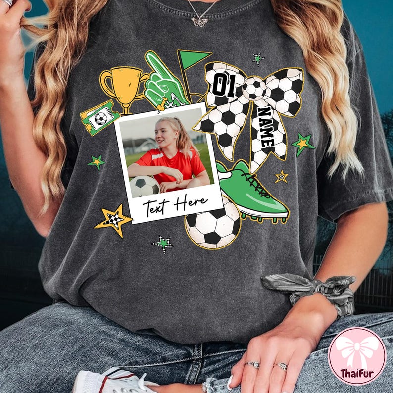 May include: A gray t-shirt with a soccer-themed design. The design includes a soccer ball, a trophy, a soccer cleat, a bow with the number "01" and the word "NAME", and a photo frame with the text "Text Here".