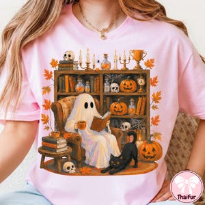 May include: A light pink t-shirt featuring a graphic of a white ghost sitting in an armchair reading a book, holding a mug, with a black cat nearby. The scene includes a bookshelf filled with books, pumpkins, candles, skulls, and autumn leaves.