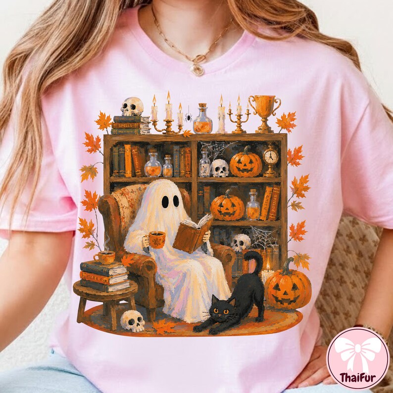 May include: A light pink t-shirt featuring a graphic of a white ghost sitting in an armchair reading a book, holding a mug, with a black cat nearby. The scene includes a bookshelf filled with books, pumpkins, candles, skulls, and autumn leaves.