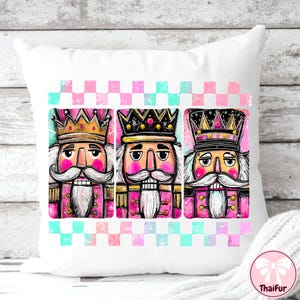 K&ouml;nnte beinhalten: Wei&szlig;es Zierkissen mit drei farbenfrohen Nussknacker-Illustrationen. Jeder Nussknacker hat ein rosa Gesicht, einen wei&szlig;en Schnurrbart und eine goldene oder schwarze Krone. Das Kissen hat einen rosa und t&uuml;rkisfarbenen karierten Rand.