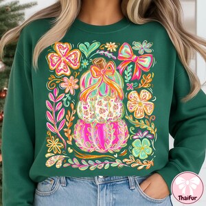 May include: Green sweatshirt featuring a stacked pumpkin design with a bow and floral accents. The pumpkins are pink, green, and white, with orange and pink bows. Surrounding the pumpkins are colorful flowers and leaves. The sweatshirt is a solid dark green color.