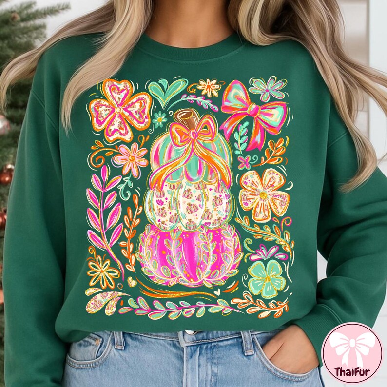 May include: Green sweatshirt featuring a stacked pumpkin design with a bow and floral accents. The pumpkins are pink, green, and white, with orange and pink bows. Surrounding the pumpkins are colorful flowers and leaves. The sweatshirt is a solid dark green color.