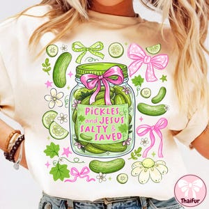 Op de afbeelding: Crèmekleurig T-shirt met een ontwerp van een pot augurken, een roze strik en de tekst "Pickles and Jesus Salty & Saved". Het ontwerp bevat groene augurken, komkommerschijfjes en decoratieve strikken.