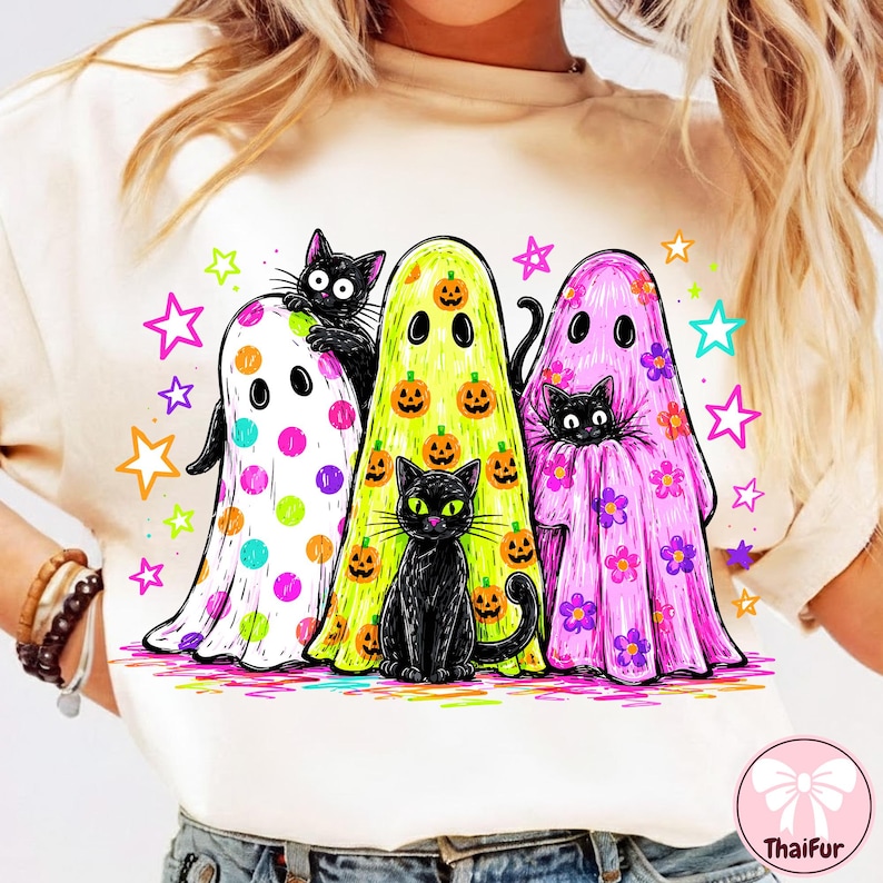 Pu&ograve; includere: Una t-shirt color crema con una grafica raffigurante tre fantasmi e due gatti neri. Un fantasma ha pois colorati, uno ha zucche arancioni e uno ha fiori viola. Stelle dai colori vivaci circondano le figure stampate sulla maglietta.