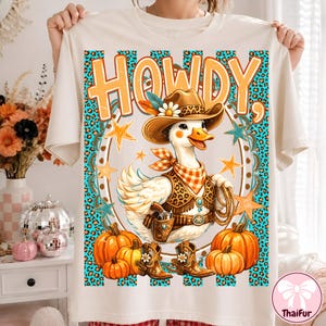 May include: A cream-colored t-shirt featuring a graphic of a duck wearing a cowboy hat, a bandana, and a vest. The duck holds a lasso and stands near pumpkins. The design includes the text "HOWDY," stars, and a turquoise leopard print background.