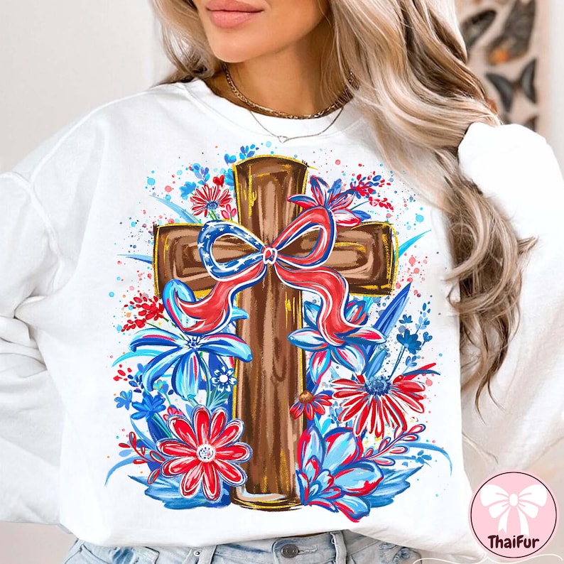 May include: White sweatshirt featuring a brown cross adorned with a red, white, and blue bow. The cross is surrounded by red and blue floral accents. The design has a watercolor effect.