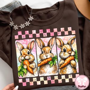 May include: A brown sweatshirt featuring three cartoon rabbits each holding a carrot. The rabbits are in rectangular frames with a pink and white checkered border. The background has a pink and white Easter egg pattern.