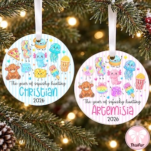 Puede incluir: Dos adornos redondos blancos con cintas, con personajes de dibujos animados coloridos, el texto "The year of squishy hunting", los nombres "CHRISTIAN" y "ARTEMISIA" y el año "2026". Uno tiene rayas azules y el otro rosas.