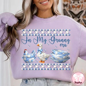 May include: A light purple sweatshirt with the text "In My Granny era". The graphic features a chicken, a goose, and a set of dishes with blue and white floral patterns. The design includes decorative borders.
