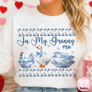 May include: White sweatshirt with a blue and white gingham and heart design. The text "In My Granny era" is written in blue script. The graphic includes a chicken, a goose, and a stack of bowls, all with a blue and white pattern.