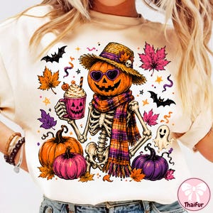 May include: A cream-colored t-shirt featuring a graphic of a skeleton wearing a straw hat, heart-shaped sunglasses, and a plaid scarf. The skeleton holds a jack-o'-lantern mug. Surrounding elements include pumpkins, bats, a ghost, and autumn leaves.