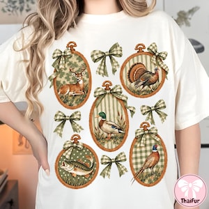 May include: An off-white t-shirt featuring a graphic print of five oval wooden embroidery hoops containing illustrations of a deer, a turkey, a mallard duck, a fish, and a pheasant, each accented with green and white checkered fabric bows.