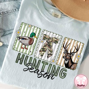 Puede incluir: Camiseta azul claro con un gr&aacute;fico de "Hunting Season". El dise&ntilde;o presenta un pato, un lazo de camuflaje y un ciervo, cada uno dentro de un marco decorativo. La camiseta tiene una etiqueta Comfort Colors.