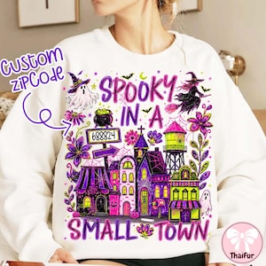 May include: A white sweatshirt featuring a colorful Halloween-themed graphic with the text "SPOOKY IN A SMALL TOWN" and "CUSTOM ZIPCODE 688824". The design includes a ghost, a witch on a broomstick, a cauldron, a water tower, and various buildings.