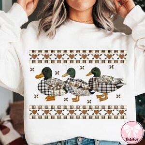 May include: White sweatshirt featuring three ducks with plaid patterned bodies and green heads. The ducks are arranged in a row below a decorative band with hearts and plaid squares. The sweatshirt has a crew neck.