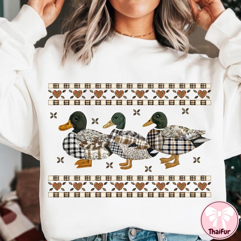 May include: White sweatshirt featuring three ducks with plaid patterned bodies and green heads. The ducks are arranged in a row below a decorative band with hearts and plaid squares. The sweatshirt has a crew neck.