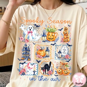 May include: A cream-colored t-shirt featuring the text Spooky Season in the air and a collage of Halloween-themed illustrations including ghosts, jack-o'-lanterns, a haunted house, a black cat, a tombstone with Trick or Treat, and floral accents.