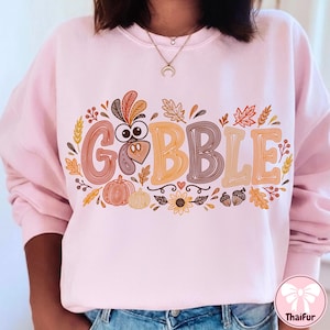 May include: A light pink long-sleeve sweatshirt featuring a graphic print with the text GOBBLE in large, stylized letters. The design incorporates a turkey face, pumpkins, autumn leaves, wheat stalks, acorns, and a sunflower in warm fall colors.