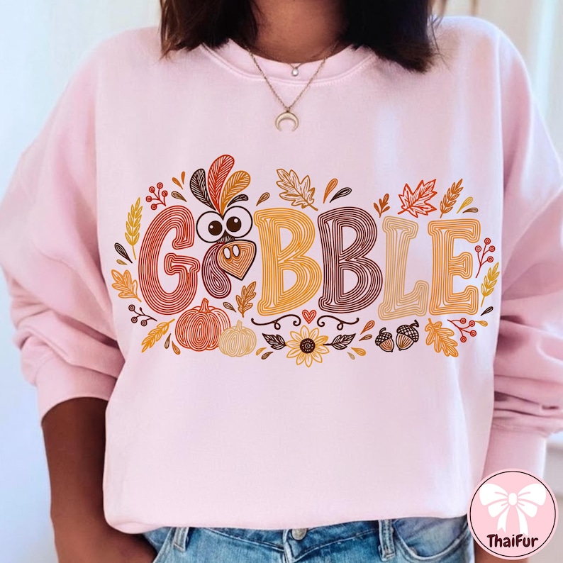 May include: A light pink long-sleeve sweatshirt featuring a graphic print with the text GOBBLE in large, stylized letters. The design incorporates a turkey face, pumpkins, autumn leaves, wheat stalks, acorns, and a sunflower in warm fall colors.