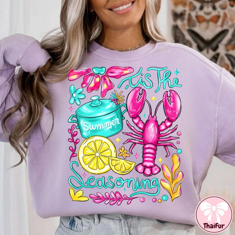 May include: A light purple sweatshirt with a colorful graphic design. The design features a pink lobster, a teal pot with the word "Summer", and the phrase "'Tis the Seasoning" in a whimsical font. Other elements include a lemon slice and floral accents.